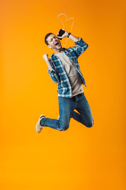 Excited young man wearing plaid shirt jumping isolated over orange background, listening to music with earphones and mobile phone - Photo, image