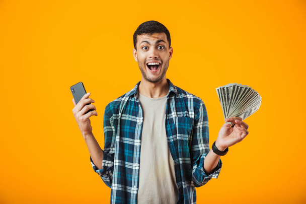 Cheerful young man wearing plaid shirt standing isolated over orange background, using mobile phone, showing money banknotes - Photo, Image