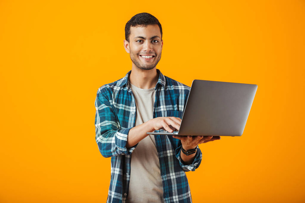 Cheerful young man wearing plaid shirt standing isolated over orange background, using laptop computer - Fotoğraf, Görsel