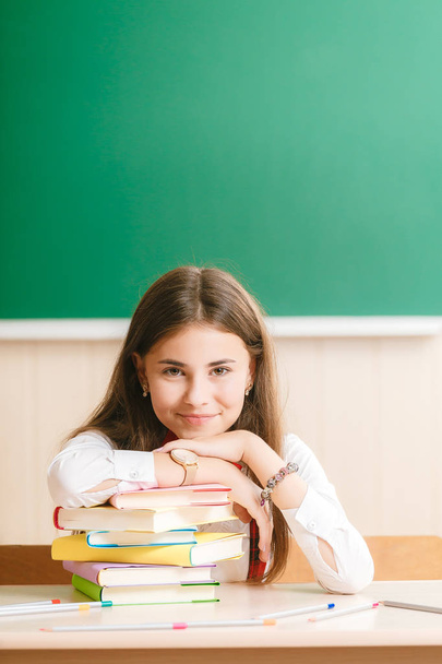 Cute brunette schoolgirl sitting at a school desk with books on the background of the blackboard. - Photo, Image