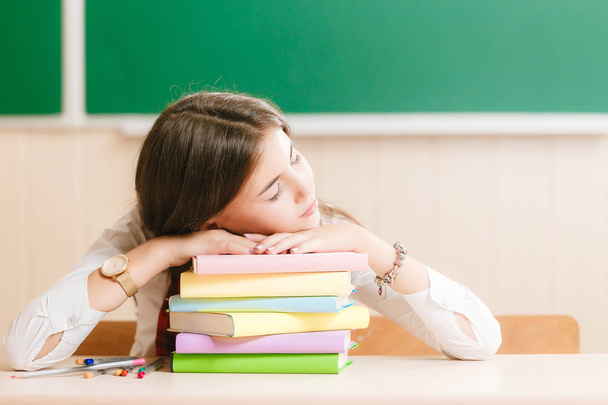 Linda colegiala morena sentada en un escritorio de la escuela con libros en el fondo de la blackboard.girl durmiendo en la mesa en los libros
 - Foto, imagen