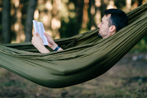 young man lying in green hammock and reading book in forest - Foto, Imagem