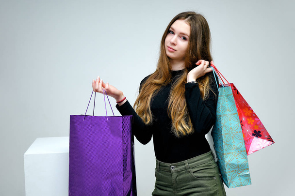 I love shopping. Portrait of a pleasant friendly and optimistic happy brunette little girl. She is in a black t-shirt smiling on a white background with packages over a white cube. - Photo, image
