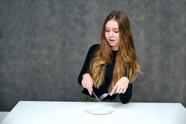 Concept Portrait of a pleasant and optimistic happy brunette girl with beautiful long hair sitting with a plate, fork and spoon. She is in a black t-shirt smiling on a white background and is eating. - Фото, изображение