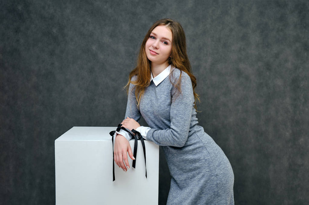 Concept portrait of a pleasant friendly manager of a happy brunette girl with beautiful long hair on a cube. She is standing in a gray dress smiling against a gray background in full growth. - Photo, Image