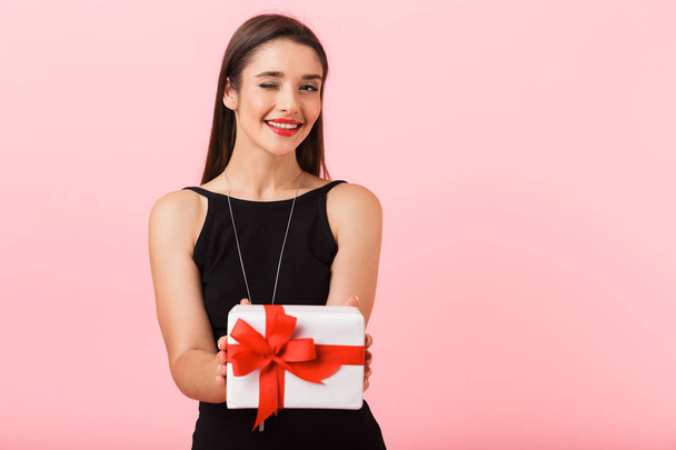 Portrait of a beautiful young woman wearing black dress standing isolated over pink background, holding gift box - Фото, изображение