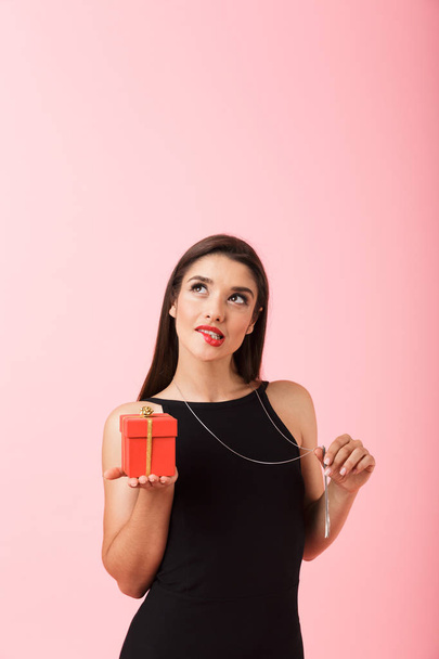 Portrait of a beautiful young woman wearing black dress standing isolated over pink background, holding gift box - Photo, image