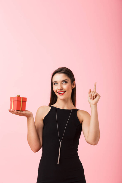 Portrait of a beautiful young woman wearing black dress standing isolated over pink background, holding gift box, pointing finger up - Foto, afbeelding