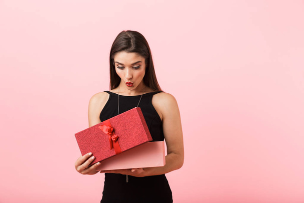 Excited woman wearing black dress holding opened gift box isolated over pink background - Foto, Imagem