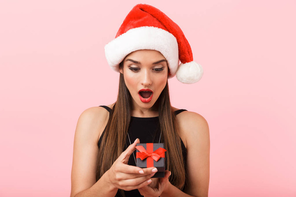 Cheerful woman wearing Christmas hat holding a gift box isolated over pink background - Photo, Image