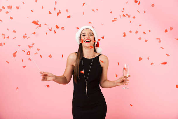 Joven alegre con sombrero de Navidad de pie aislado sobre fondo rosa, sosteniendo una copa de champán espumoso, celebrando
 - Foto, imagen