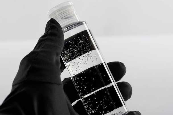 vista recortada de la mano femenina en guante negro con botella cosmética con líquido transparente sobre fondo blanco
 - Foto, Imagen