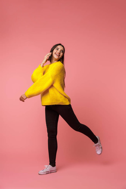 Full length of a cheerful young girl wearing sweater standing isolated over pink background, talking on mobile phone - Foto, afbeelding