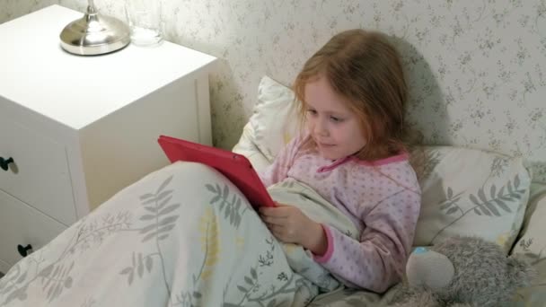 little girl in bed playing on tablet - Materiał filmowy, wideo
