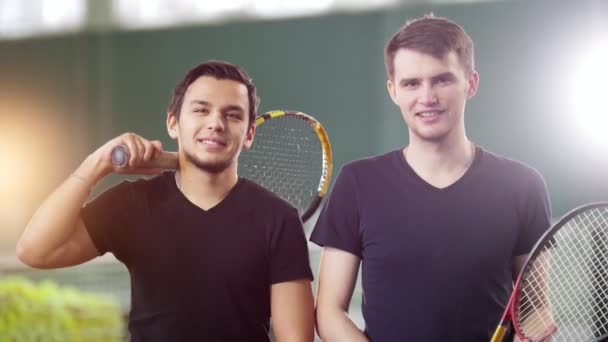 Szkolenia na kort tenisowy. Dwóch młodych mężczyzn, patrząc w kamerę, trzymając rakiety - Materiał filmowy, wideo