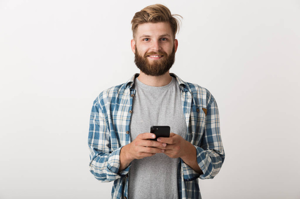 Photo of a handsome young bearded man standing isolated over white wall background using mobile phone. - Фото, зображення