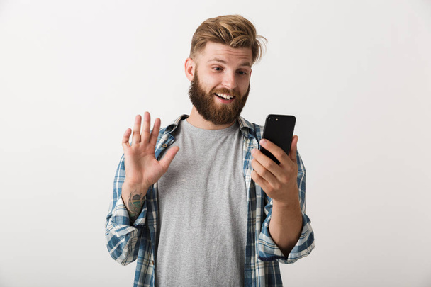 A csinos, fiatal, szakállas férfi áll elszigetelt használ mozgatható telefon beszélő integetett fehér háttér fénykép. - Fotó, kép