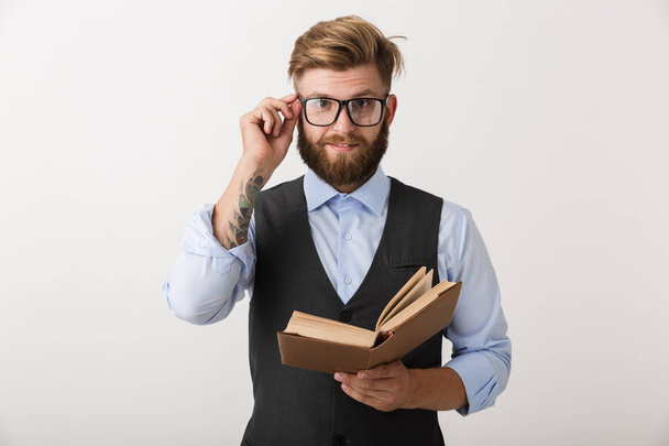 Image of a handsome young bearded man standing isolated over white wall background holding book reading. - Foto, imagen