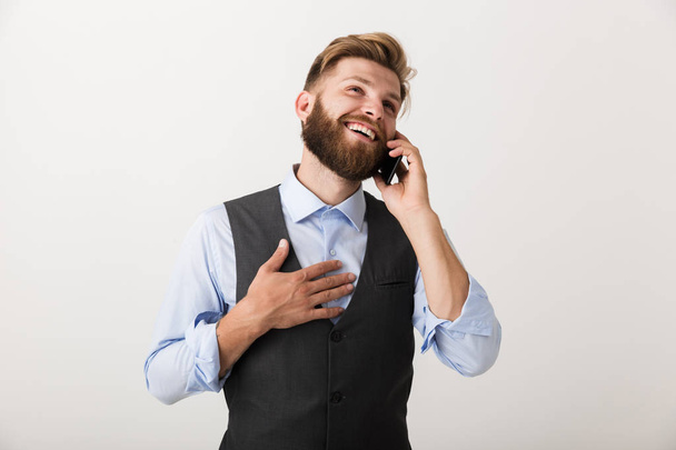 Picture of a handsome young bearded man standing isolated over white wall background talking by mobile phone. - Φωτογραφία, εικόνα