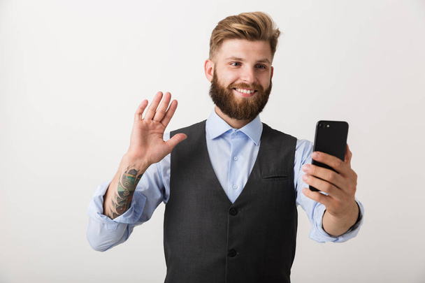 Yakışıklı bir genç sakallı adam cep telefonu konuşan sallayarak kullanarak beyaz duvar arka plan üzerinde izole ayakta fotoğrafı. - Fotoğraf, Görsel