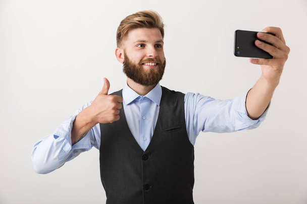 Picture of a handsome young bearded man standing isolated over white wall background take a selfie by mobile phone. - Valokuva, kuva