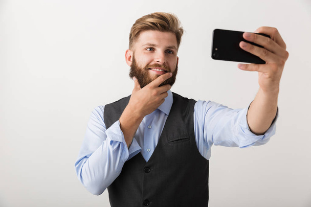 Obrázek pohledný mladý vousatý muž stojící izolované nad bílou stěnu pozadí focení mobilem. - Fotografie, Obrázek