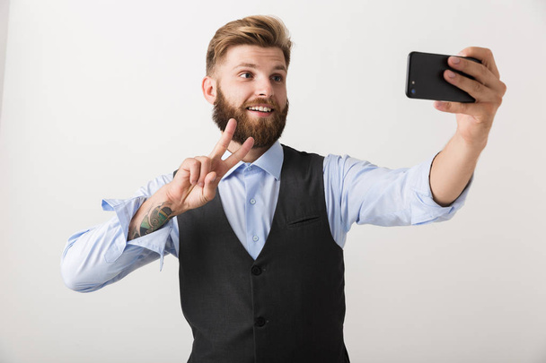 Picture of a handsome young bearded man standing isolated over white wall background take a selfie by mobile phone. - Photo, Image