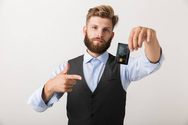Photo of a handsome young bearded man standing isolated over white wall background holding credit card pointing. - Φωτογραφία, εικόνα