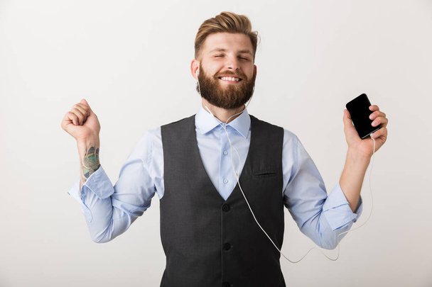 Photo of a handsome young bearded man standing isolated over white wall background holding mobile phone listening music. - Foto, imagen