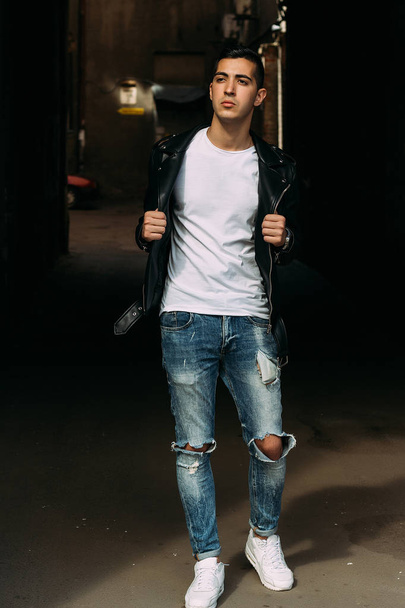 young, handsome guy in a white T-shirt, black, leather jacket and blue, torn jeans is on the street, model, city walk, live style.in white sneakers - Φωτογραφία, εικόνα