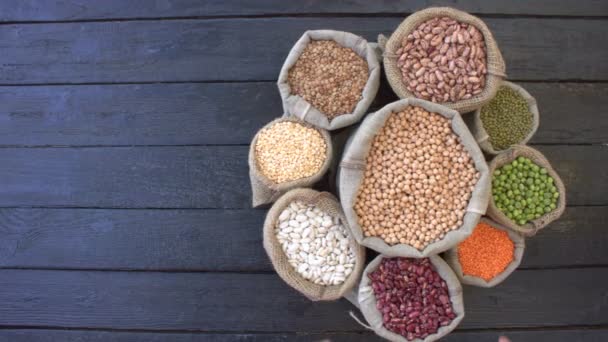 Legumes on dark wooden ecological background.Beans are located in the shape of circle on black wooden table. Hands take chickpeas and pour back. Bean cultures in knitted bags. Camera moves from left to right.  - Footage, Video
