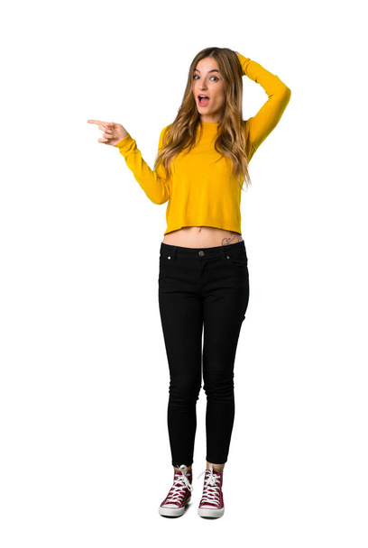A full-length shot of a young girl with yellow sweater pointing finger to the side and presenting a product on isolated white background - Photo, Image