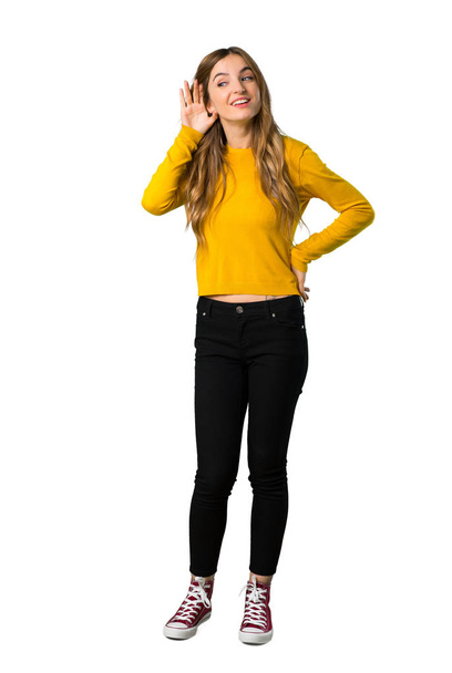 A full-length shot of a young girl with yellow sweater listening to something by putting hand on the ear on isolated white background - Фото, изображение