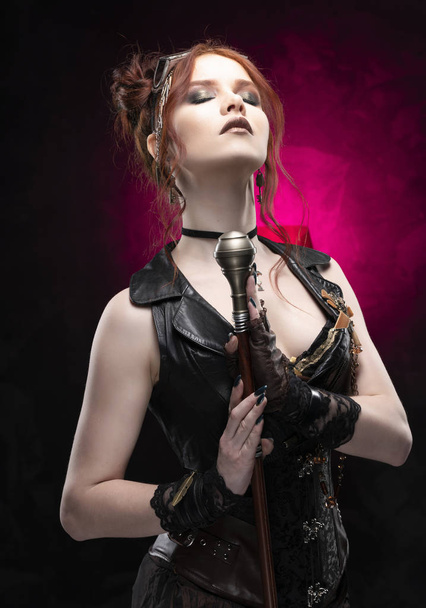 A beautiful red-haired cosplayer girl wearing a Victorian-style steampunk costume with large breasts in a deep neckline and glasses holding a cane on a black and purple background. Copy space. - Fotoğraf, Görsel
