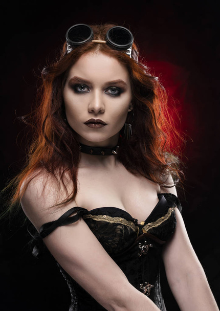 Beautiful smiling redhead cosplayer girl wearing a Victorian-style steampunk costume with a big breast in a deep neckline on red and black background. Portrait. Copy space. - Φωτογραφία, εικόνα