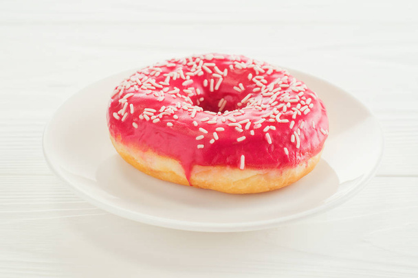 Un donut rosa, de cerca. Donut en un plato sobre una mesa de madera blanca. Delicioso desayuno, rosquilla dulce con espacio para copiar
.  - Foto, imagen