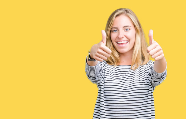 Beautiful young woman wearing stripes sweater over isolated background approving doing positive gesture with hand, thumbs up smiling and happy for success. Looking at the camera, winner gesture. - Photo, Image