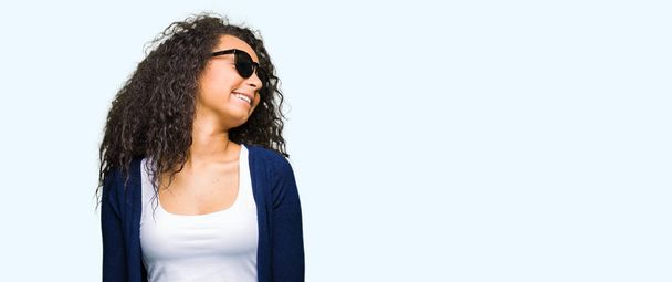 Young beautiful girl with curly hair wearing fashion sunglasses looking away to side with smile on face, natural expression. Laughing confident. - Photo, image