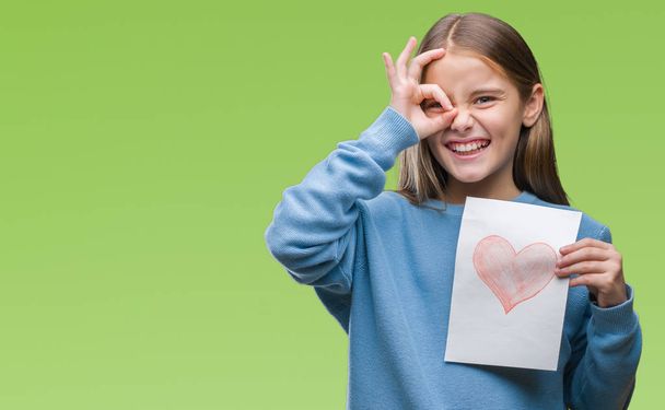 Young beautiful girl giving mother father day card with red heart over isolated background with happy face smiling doing ok sign with hand on eye looking through fingers - Photo, Image