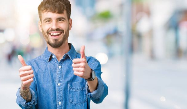 Young handsome man over isolated background approving doing positive gesture with hand, thumbs up smiling and happy for success. Looking at the camera, winner gesture. - Photo, Image
