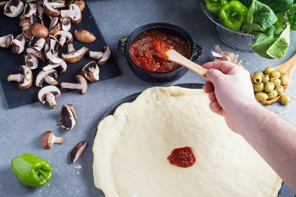 Pizza preparation, raw dough surrounded by ingredients. Male hand smudging sauce on pizza dough.  - Photo, Image