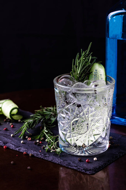 Gin-tonic alcoholic cocktail. Cucumber, rosemary, ice. On a black wooden background. - Foto, Imagen