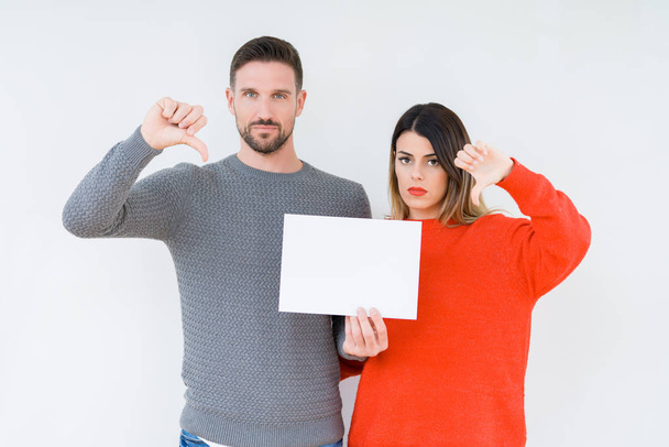 Young couple holding blank sheet paper over isolated background with angry face, negative sign showing dislike with thumbs down, rejection concept - Foto, afbeelding