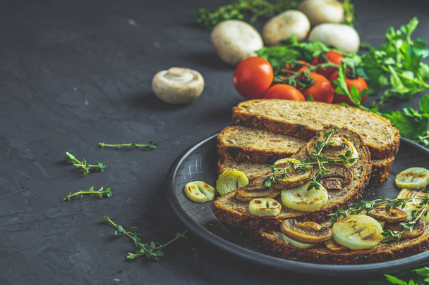Delicious homemade whole grain rye sliced bread toast with fried cutting mushroom champignon and onion on black ceramic plate, herbs and vegatables, dark concrete table surface, copy space. - Photo, image