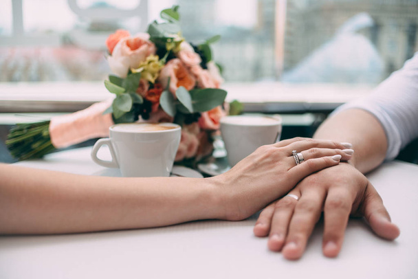 Жених и невеста держатся за руки, держа дорогие обручальные кольца с белым золотом, на фоне свадебного букета и двух чашек кофе на белом столе
 - Фото, изображение