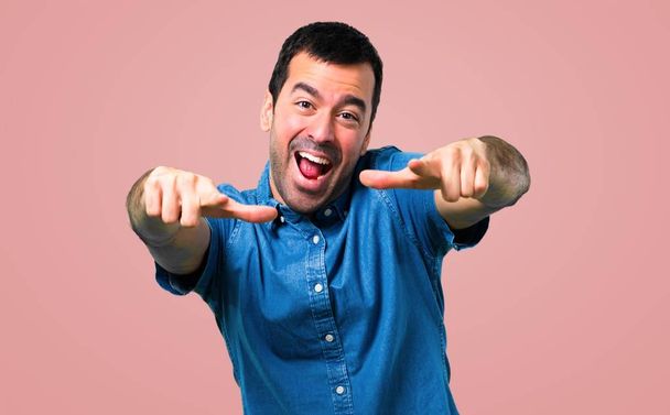Handsome man with blue shirt pointing with finger at someone and laughing a lot on pink background - Photo, Image