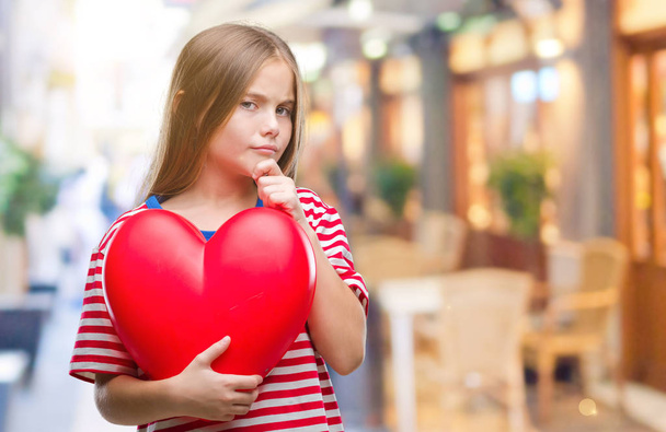 Mooi meisje rood hart houdt over geïsoleerde achtergrond ernstige gezicht na te denken over de vraag, zeer verward idee - Foto, afbeelding