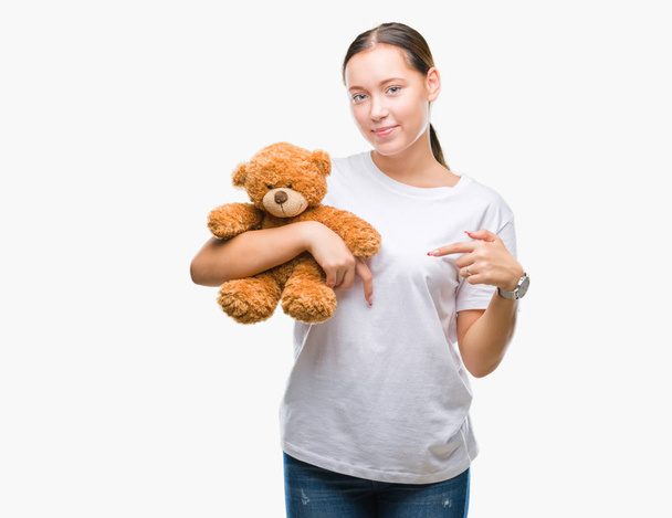 Молода біла жінка тримає плюшевого ведмедя над ізольованим тлом з несподіваним обличчям, спрямованим пальцем до себе
 - Фото, зображення