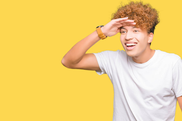 Young handsome man with afro hair wearing casual white t-shirt very happy and smiling looking far away with hand over head. Searching concept. - Photo, Image