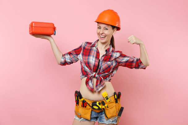 Strong young woman in orange helmet, plaid shirt, denim shorts, kit tools belt full of instruments, toolbox showing biceps, muscles isolated on pink background. Female male work. Renovation concept - Photo, Image